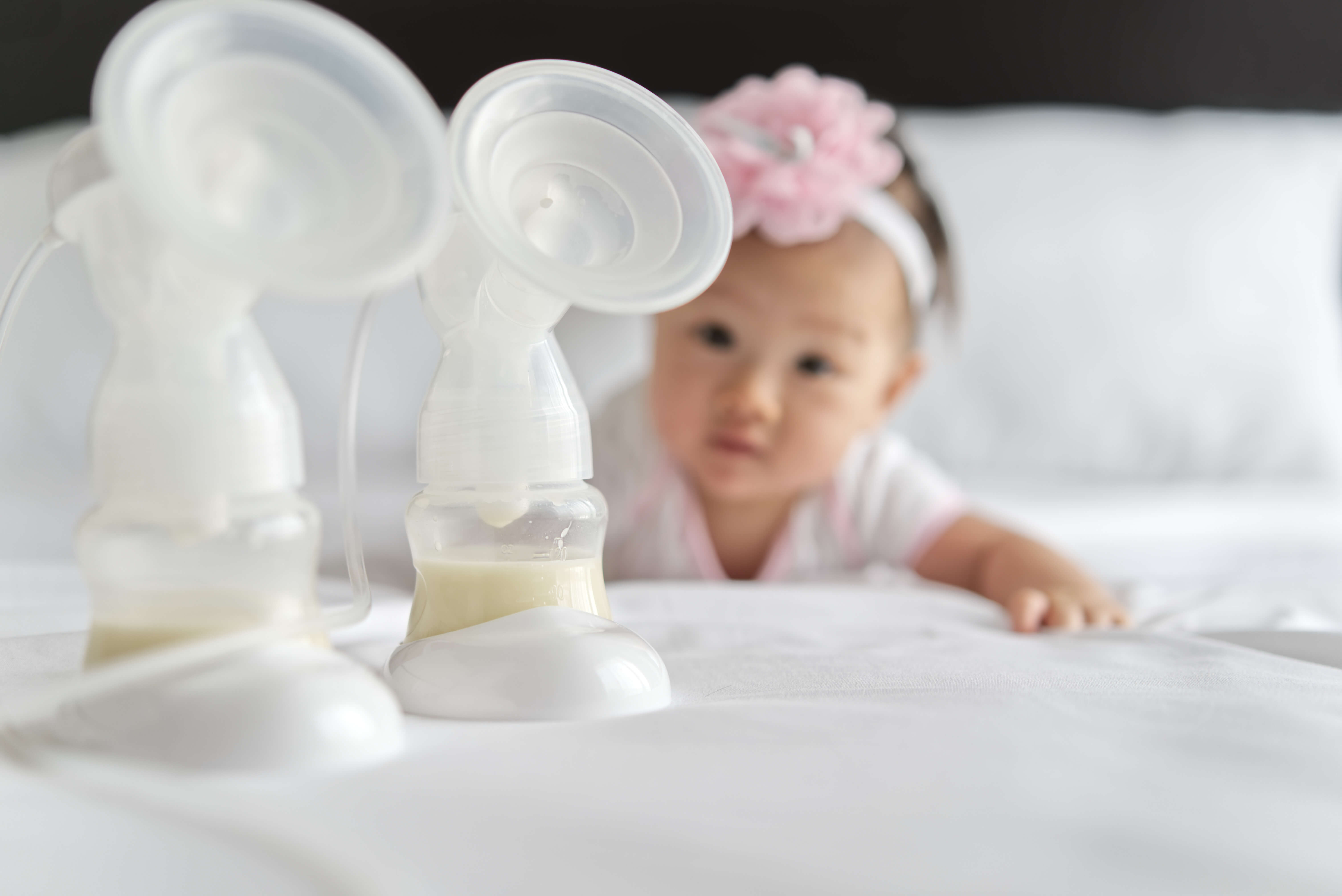 Breast milk in milk pump's bottles on the bed with little cute baby crawling in background. The milk got from the pump's machine and ready for the baby. Focus on milk bottle.