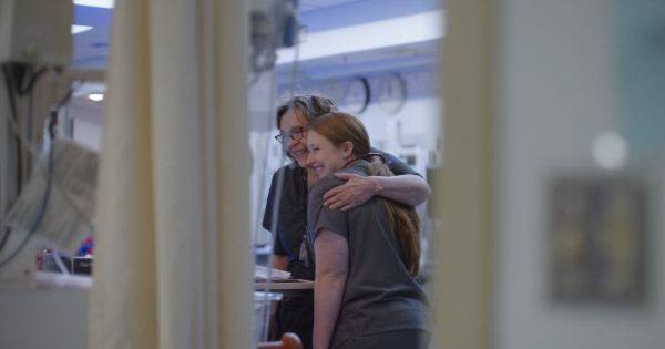 Two nurses share a supportive embrace inside a hospital patient care area.