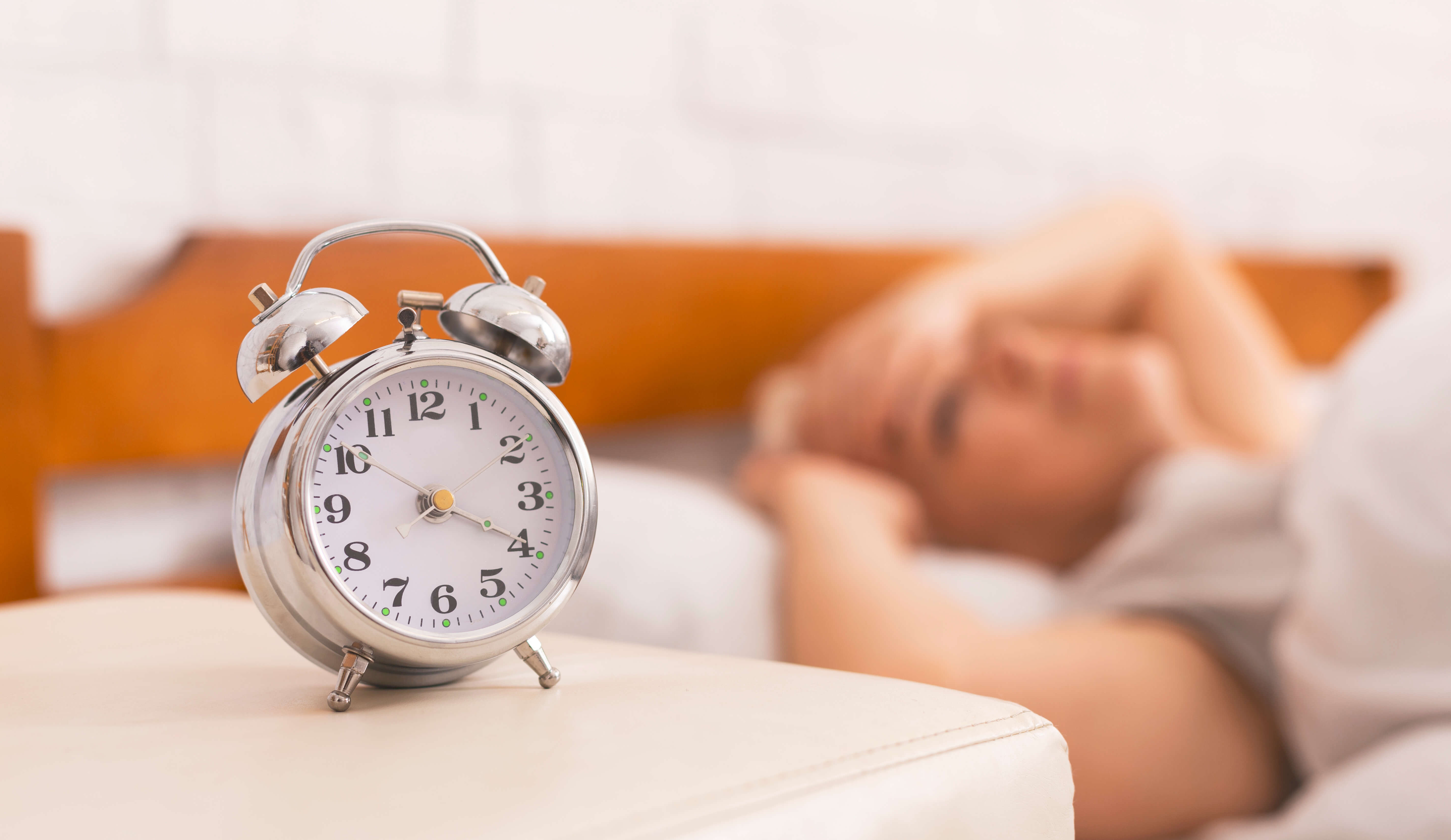 Alarm clock in foreground as a woman lies awake in bed, struggling with sleeplessness.