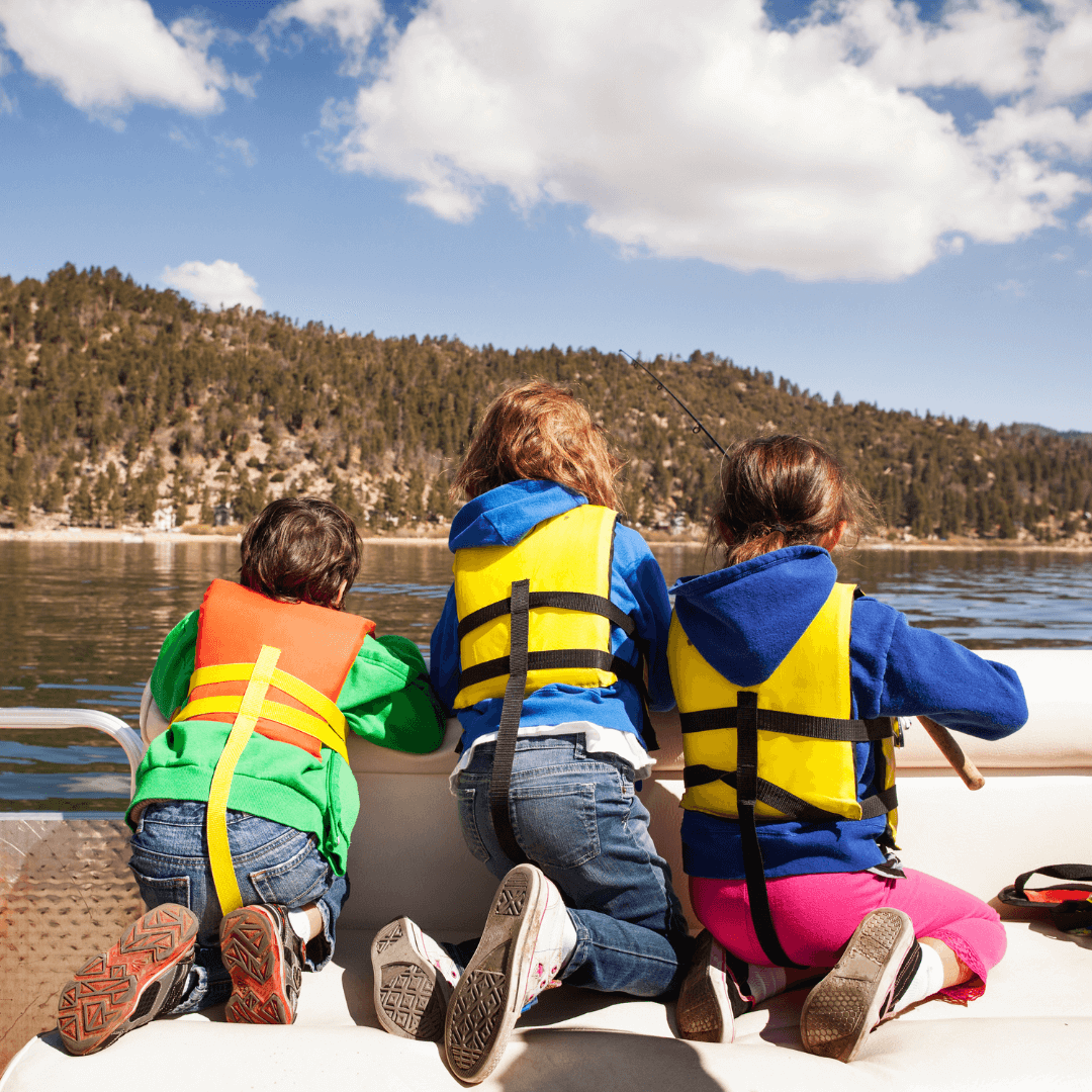 Three children on a boat looking at the lake with there life jackets on.