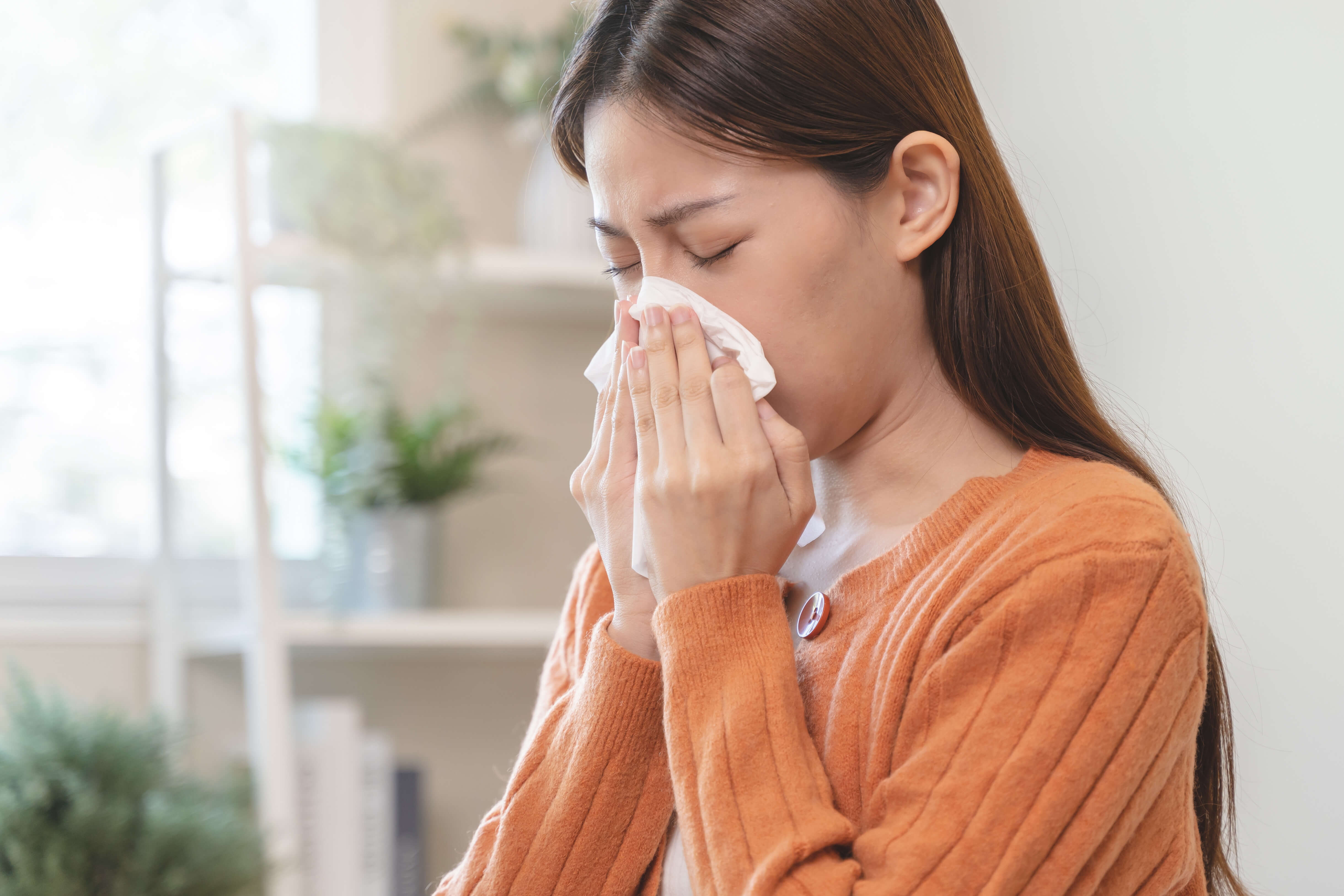 An asian woman blowing her nose into a tissue as she is struggling with pollen allergies.