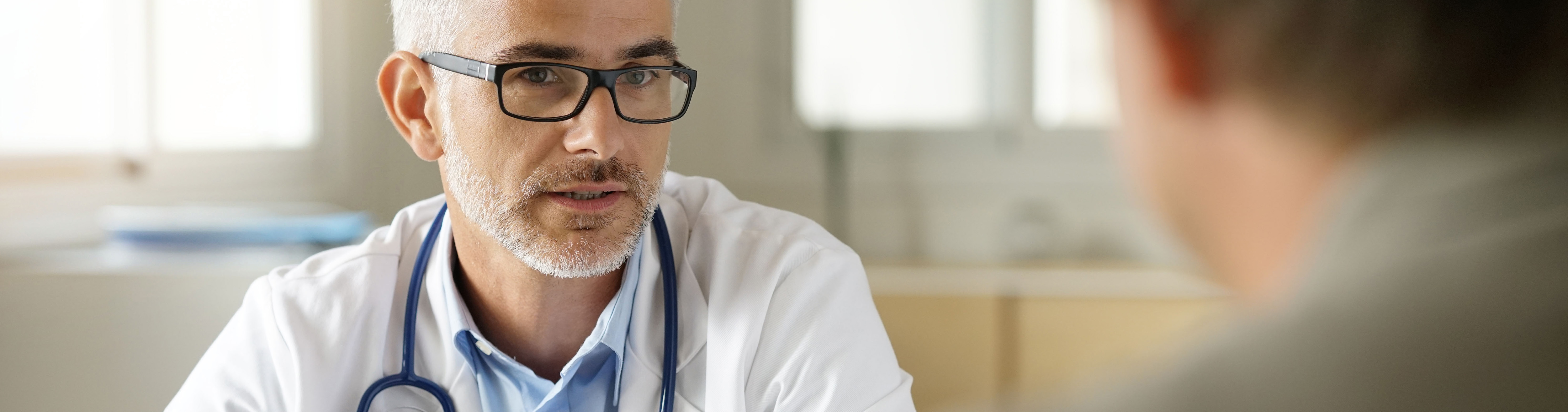 Provider dressed in a white coat wearing glasses and a stethoscope listening to a patient.